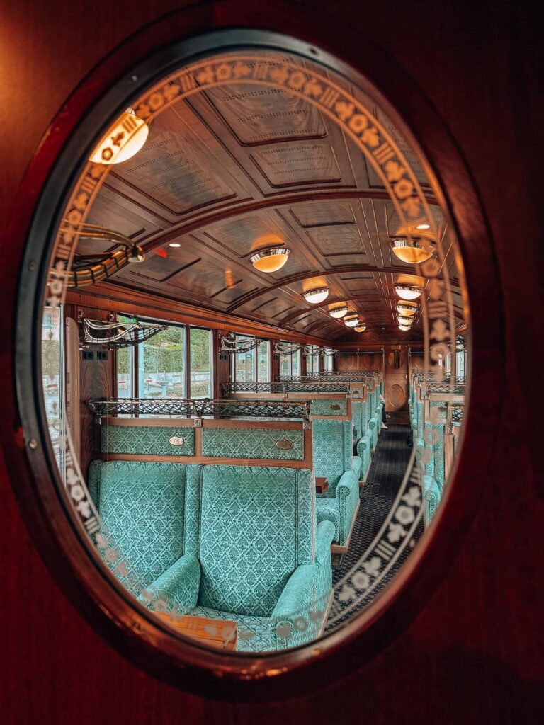 A view through an oval glass window into the GoldenPass Belle Epoque train’s first-class cabin, showing rows of elegant green patterned seats, ornate wood paneling, and warm vintage lighting that captures the train’s timeless charm.