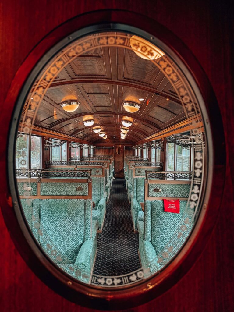 View through an oval glass window into the GoldenPass Belle Epoque train’s first-class carriage, revealing rows of elegant green velvet seats, ornate wood paneling, and warm vintage lighting that evoke the charm of early 1900s train travel.
