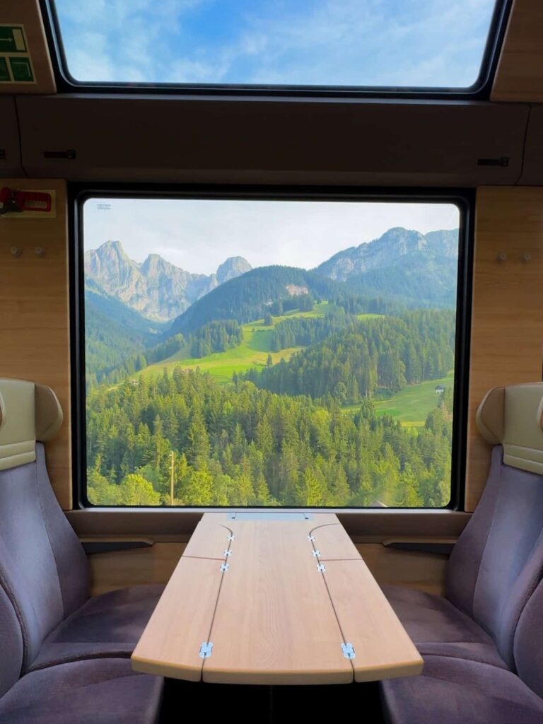 Scenic view from a modern GoldenPass panoramic train with large windows and sleek beige seats, framing a breathtaking landscape of green hills, pine forests, and distant alpine peaks under a bright blue sky.