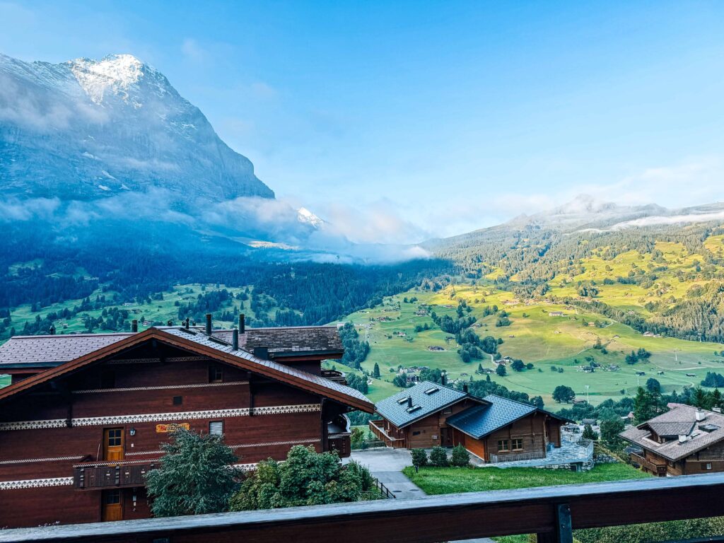 Morning view over Grindelwald village with traditional wooden chalets, lush green valley, and snow-capped mountains partially covered by clouds under a clear blue sky in the Swiss Alps.