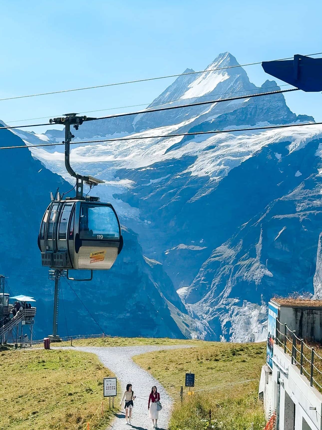 Cable car ascending toward the snow-capped mountains at Grindelwald First in Switzerland, with two women walking along a scenic alpine path below.