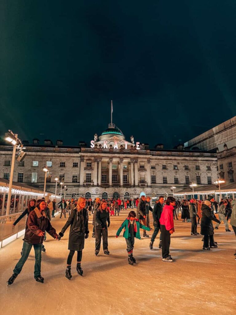 People ice skating under the night sky at Somerset House in London, surrounded by warm lights and festive decorations with the grand building facade in the background.