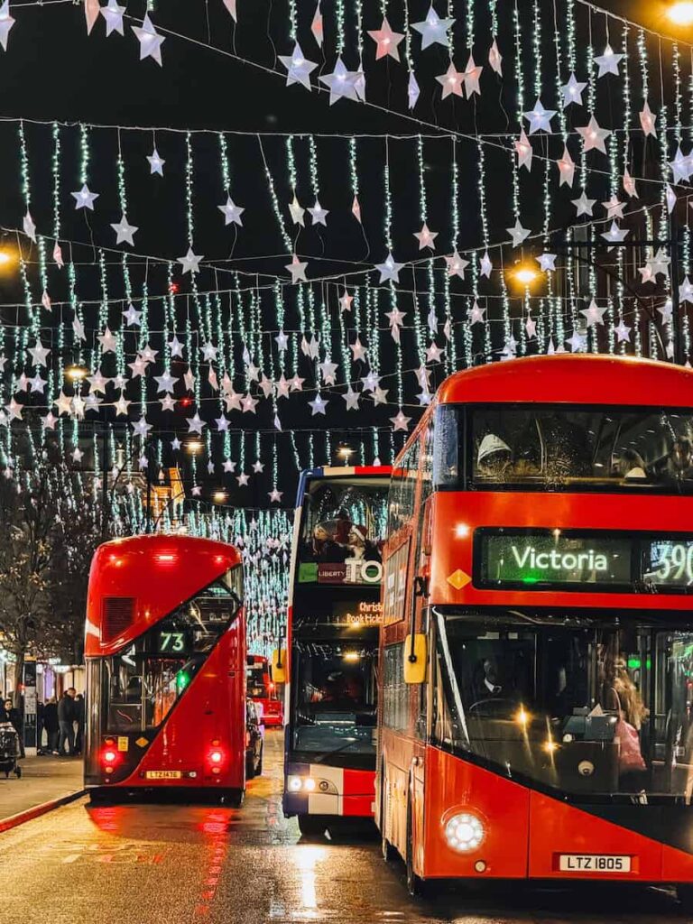 Classic red London double-decker buses driving through a street lined with twinkling star-shaped Christmas lights hanging overhead during the festive season.
