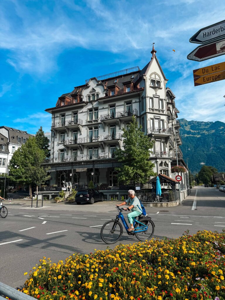 Elegant historic building in Interlaken, Switzerland, with balconies and mountain views, as a cyclist rides past colorful flower beds under a bright blue sky.