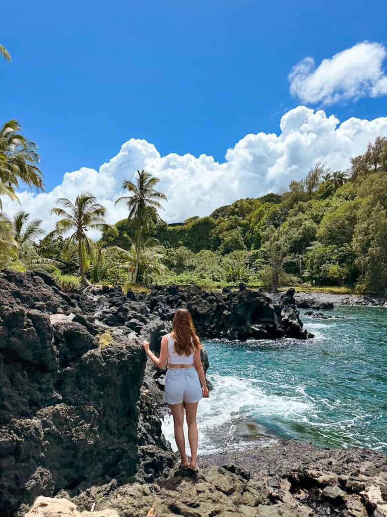 Woman standing on black lava rocks overlooking the turquoise ocean and lush green cliffs at Ke‘anae Peninsula on Maui’s Road to Hana