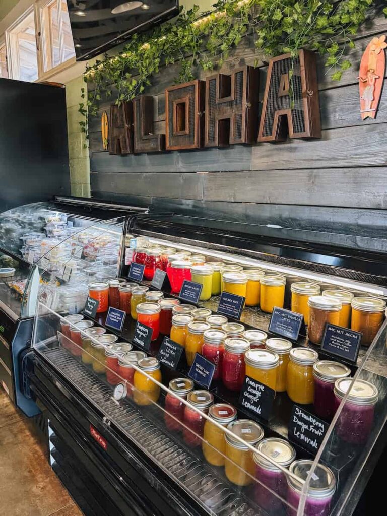 Display of fresh juices and coffee drinks in glass jars inside Kuau Store in Paia, Maui, with a wooden ‘Aloha’ sign above the counter