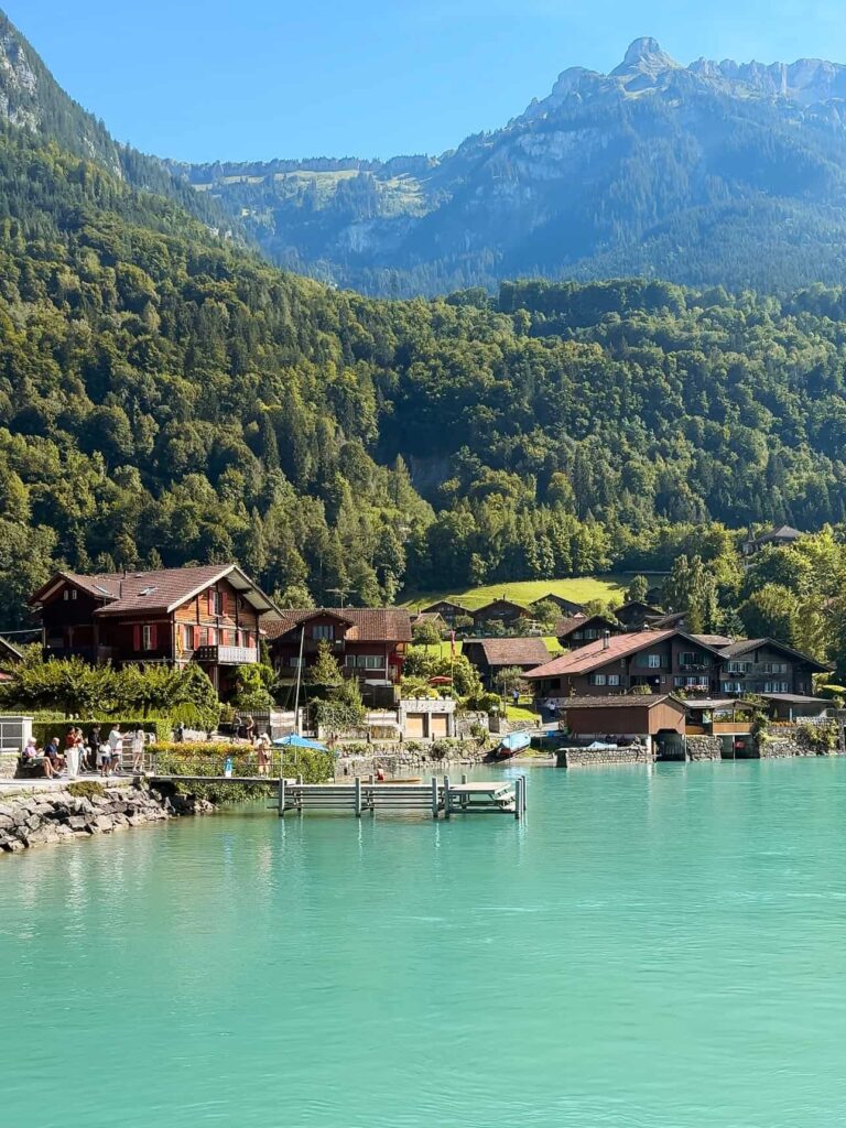 Peaceful lakeside view of wooden chalets along the turquoise waters of Lake Brienz, framed by forested mountains under a clear blue sky.