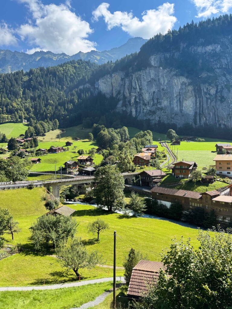 Panoramic view of Lauterbrunnen Valley with lush green meadows, alpine chalets, a river, and steep rocky cliffs surrounded by forested mountains under a bright blue sky.