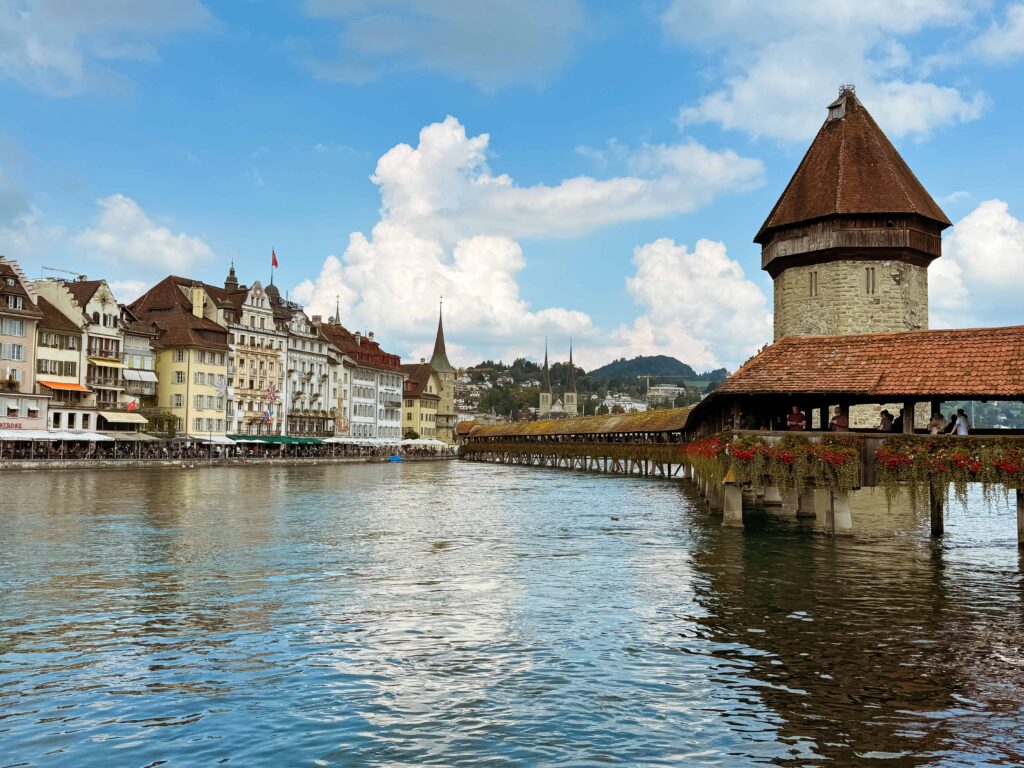 View of Lucerne’s iconic Chapel Bridge and Water Tower reflected in the Reuss River, surrounded by colorful historic buildings and distant church spires under a bright blue sky with scattered clouds.