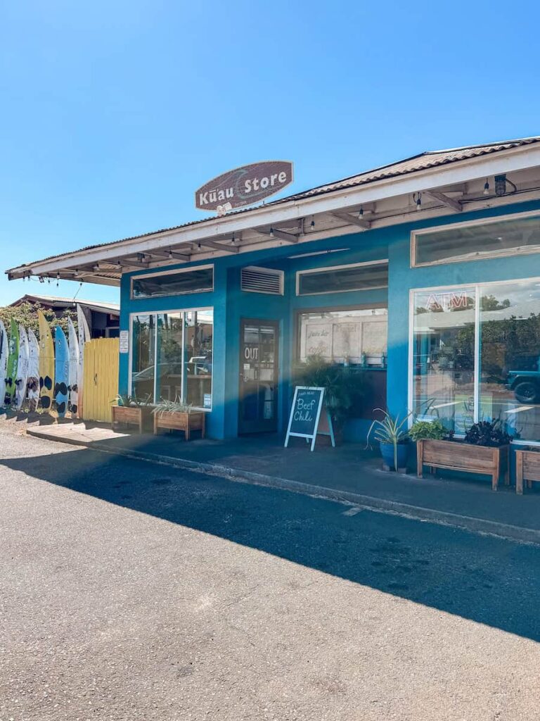 Front view of the bright turquoise Kuau Store in Paia, Maui, with a sign above the entrance and surfboards displayed along the side