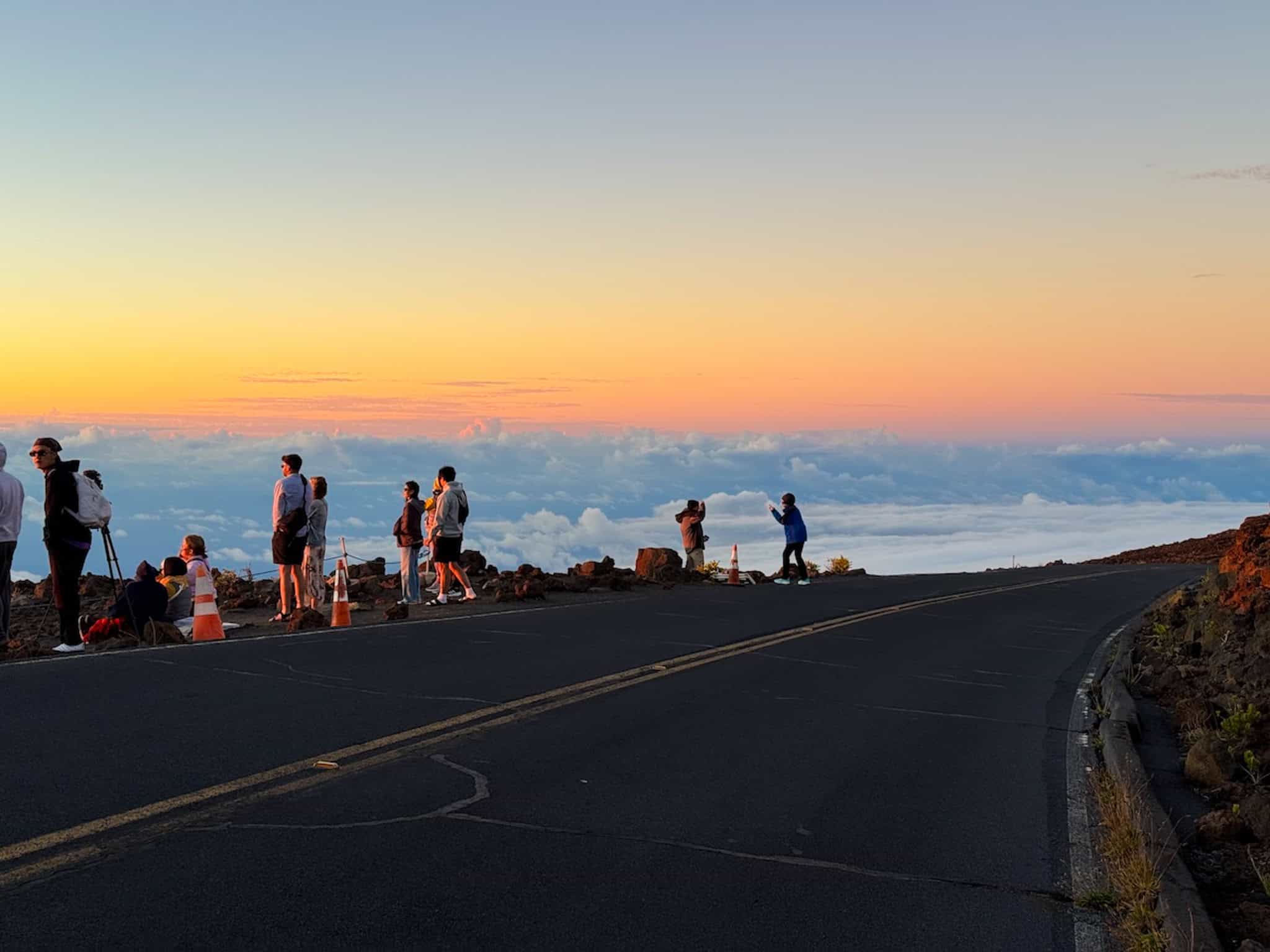Visitors gathered along the road at Haleakalā National Park to watch the sunset above the clouds, with a glowing orange and pink sky.