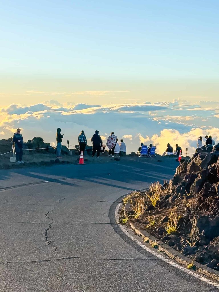 Visitors standing along the road at Haleakalā Summit watching the sunset above a sea of clouds in Maui.