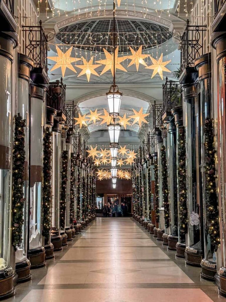 The Piccadilly Arcade in London adorned with elegant Christmas decorations, featuring glowing star-shaped lights hanging from the ceiling and garlands wrapped around the shop columns.