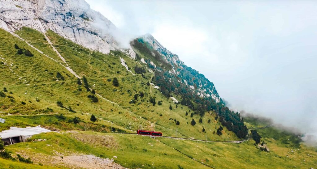 Bright red cogwheel train ascending the steep green slopes of Mount Pilatus in Switzerland, surrounded by misty clouds and rugged alpine cliffs.