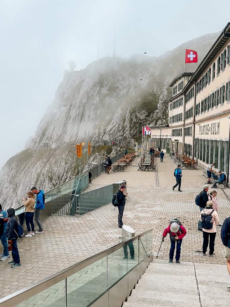 Visitors walking and taking photos on the viewing terrace at Pilatus Kulm, Switzerland, surrounded by mist-covered mountain peaks and buildings flying Swiss flags.