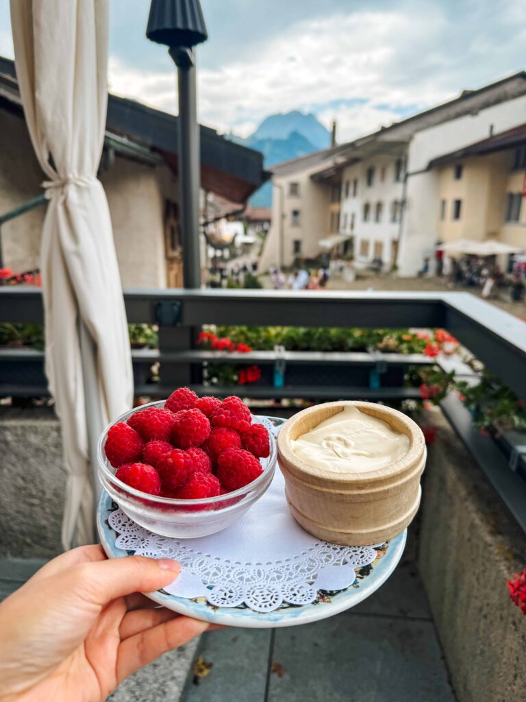 Bowl of fresh raspberries and wooden cup of double cream held over a balcony with a view of Gruyères village and surrounding mountains.