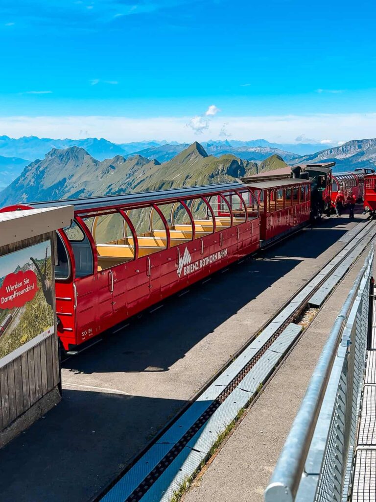 Red Brienz Rothorn Bahn train parked at the Rothorn Kulm station platform with sweeping mountain views and bright blue skies above.