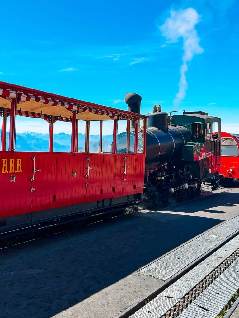 Classic Brienz Rothorn Bahn steam train with red wooden carriages and a black locomotive at the summit station with mountain peaks in the background.