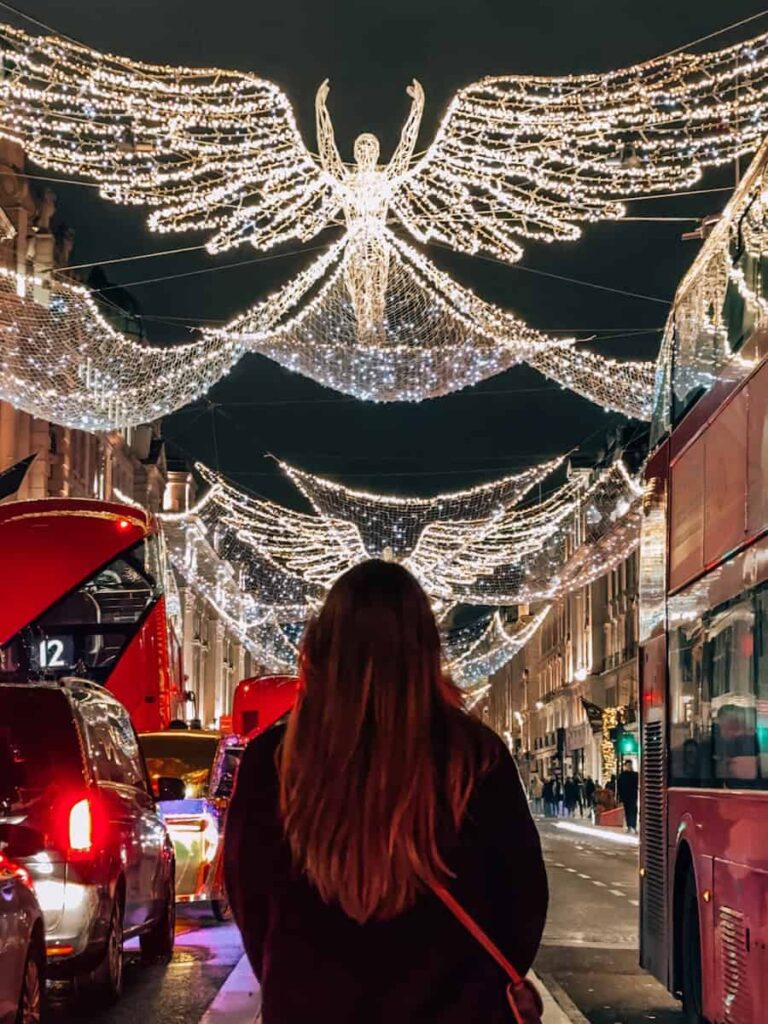 A person standing on Regent Street in London, gazing at the illuminated angel light displays that stretch above the road, with red buses and taxis passing by beneath the twinkling holiday decorations.