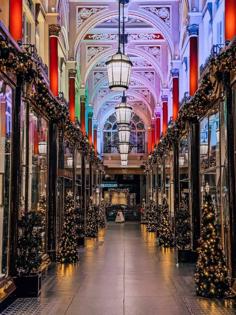The Royal Arcade in London decorated for Christmas with twinkling trees lining the walkway, grand hanging lanterns, and colorful lights illuminating the ornate arched ceiling.