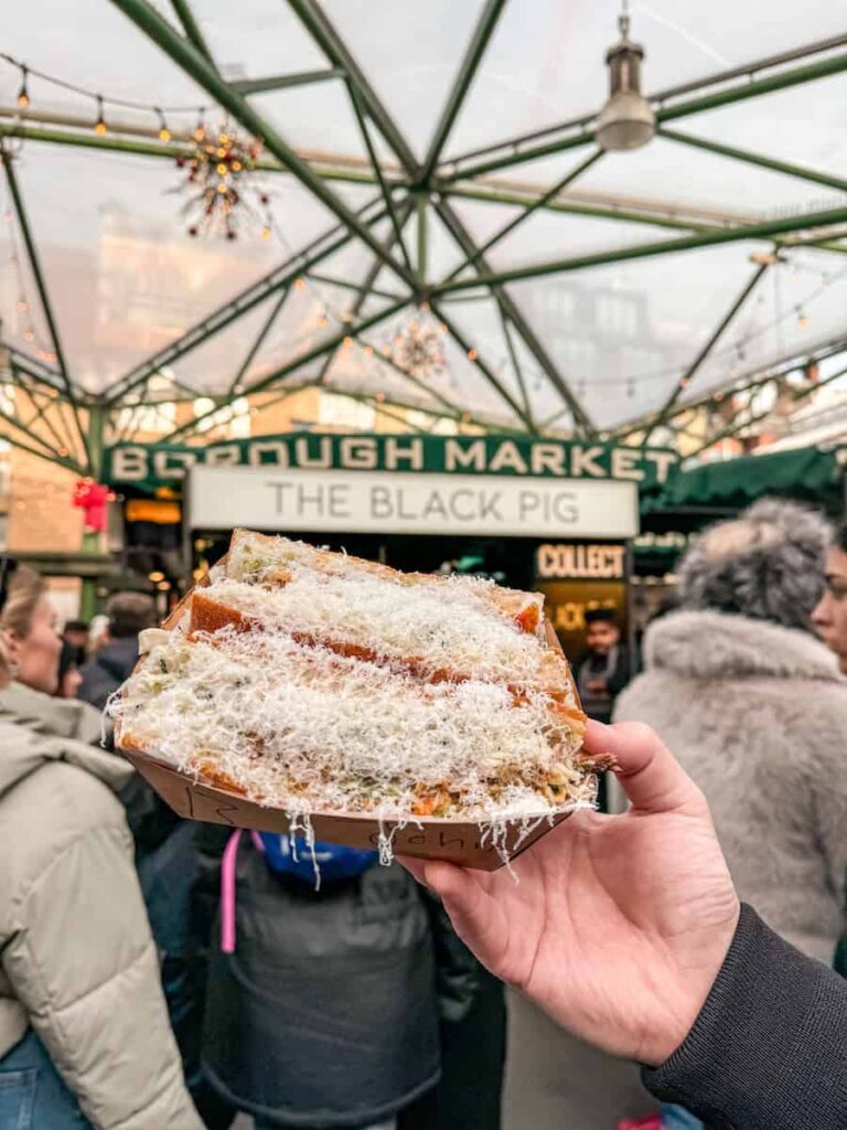 A close-up of a hand holding a sandwich covered in shaved cheese from The Black Pig stall at Borough Market, surrounded by people and festive market lights.