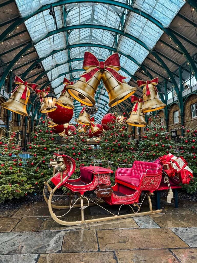 A festive Christmas display inside Covent Garden Market in London featuring a red sleigh surrounded by Christmas trees decorated with red ornaments and oversized golden bells hanging from the glass ceiling.