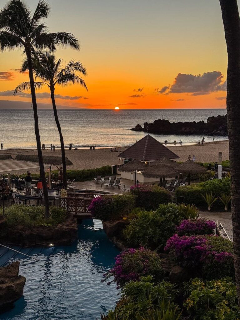 Vibrant orange sunset over Kaʻanapali Beach and Black Rock at Sheraton Maui Resort, with palm trees and the ocean reflecting golden light.
