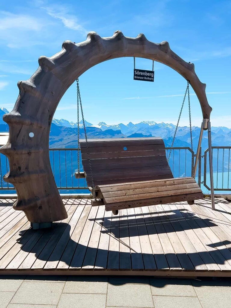 Wooden swing structure labeled “Sörenberg Brienzer Rothorn” overlooking snow-capped Swiss Alps and turquoise Lake Brienz on a sunny day.