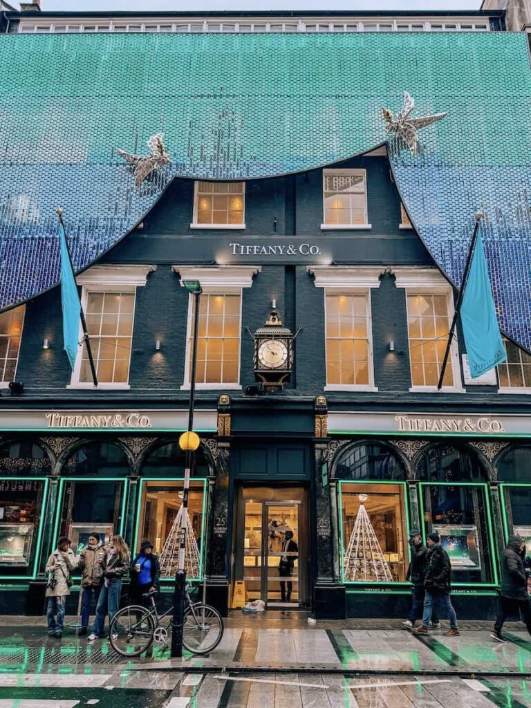 The exterior of the Chanel boutique in London decorated for Christmas with a massive black and gold bow made of tweed fabric draped across the building facade, glowing lights accenting the elegant holiday display.