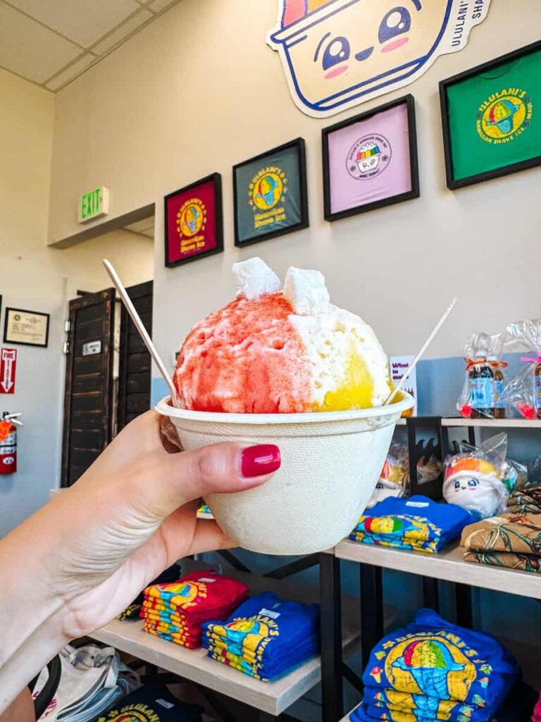 Hand holding a colorful bowl of Ululani’s Hawaiian Shave Ice inside the shop, with bright logo signs and merchandise in the background.