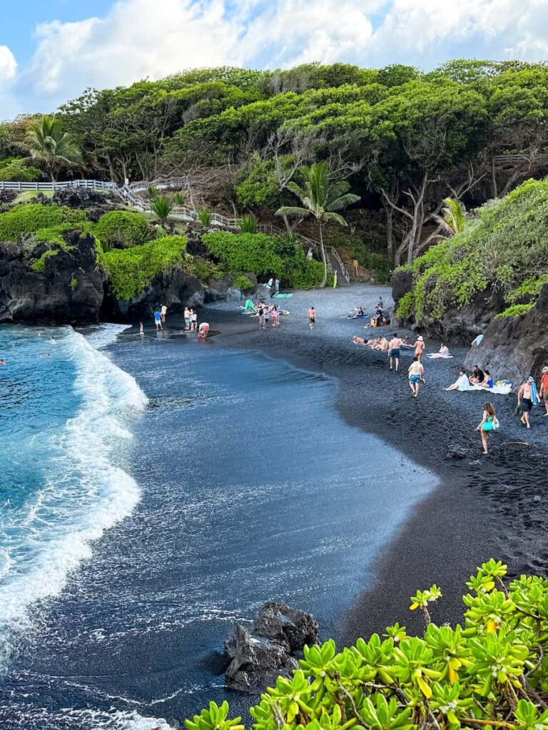View of the famous black sand beach at Waiʻanapanapa State Park on the Road to Hana, surrounded by lush green cliffs and turquoise waves. A must see during a 5 day Maui itinerary. 