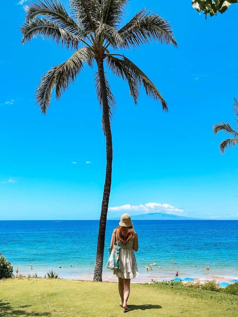 Woman in a summer dress and sun hat standing under a palm tree overlooking Wailea Beach in Maui, with turquoise water and beachgoers below