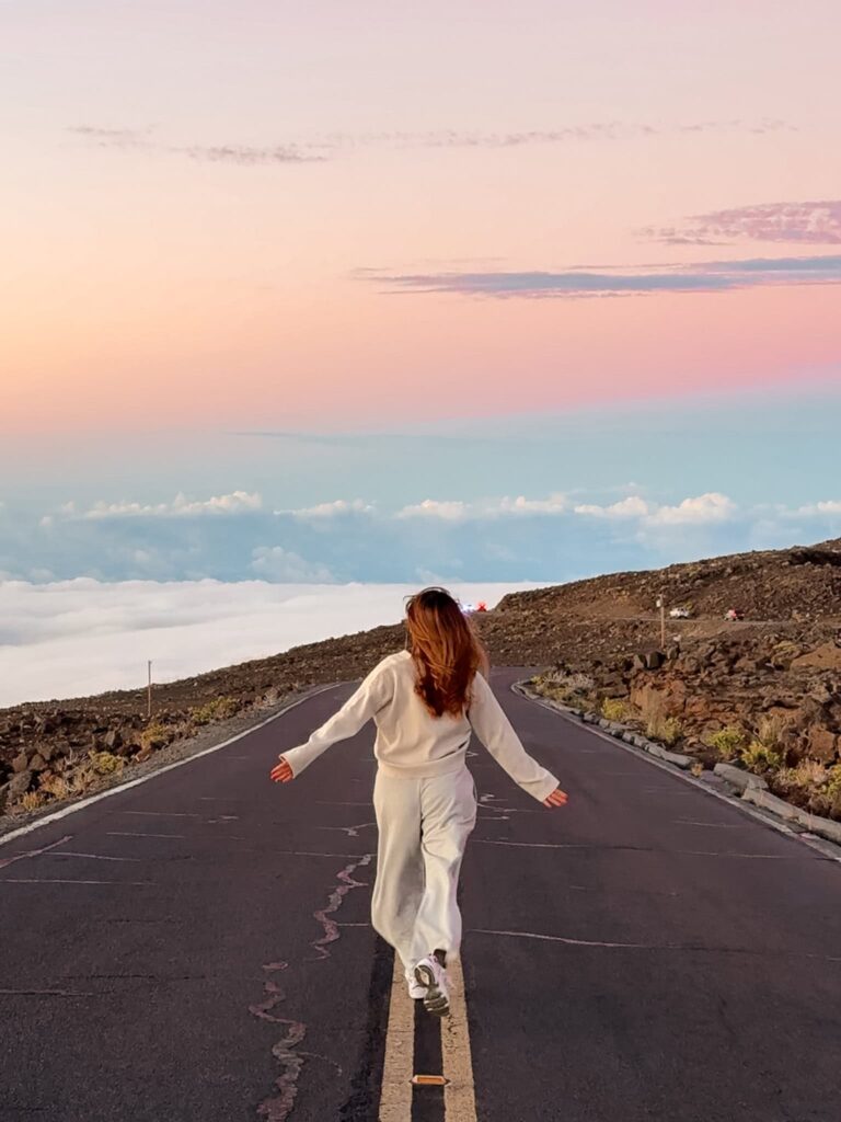 A woman walking along the road at Haleakalā Summit at sunset, surrounded by rocky slopes and pastel clouds glowing pink and blue.