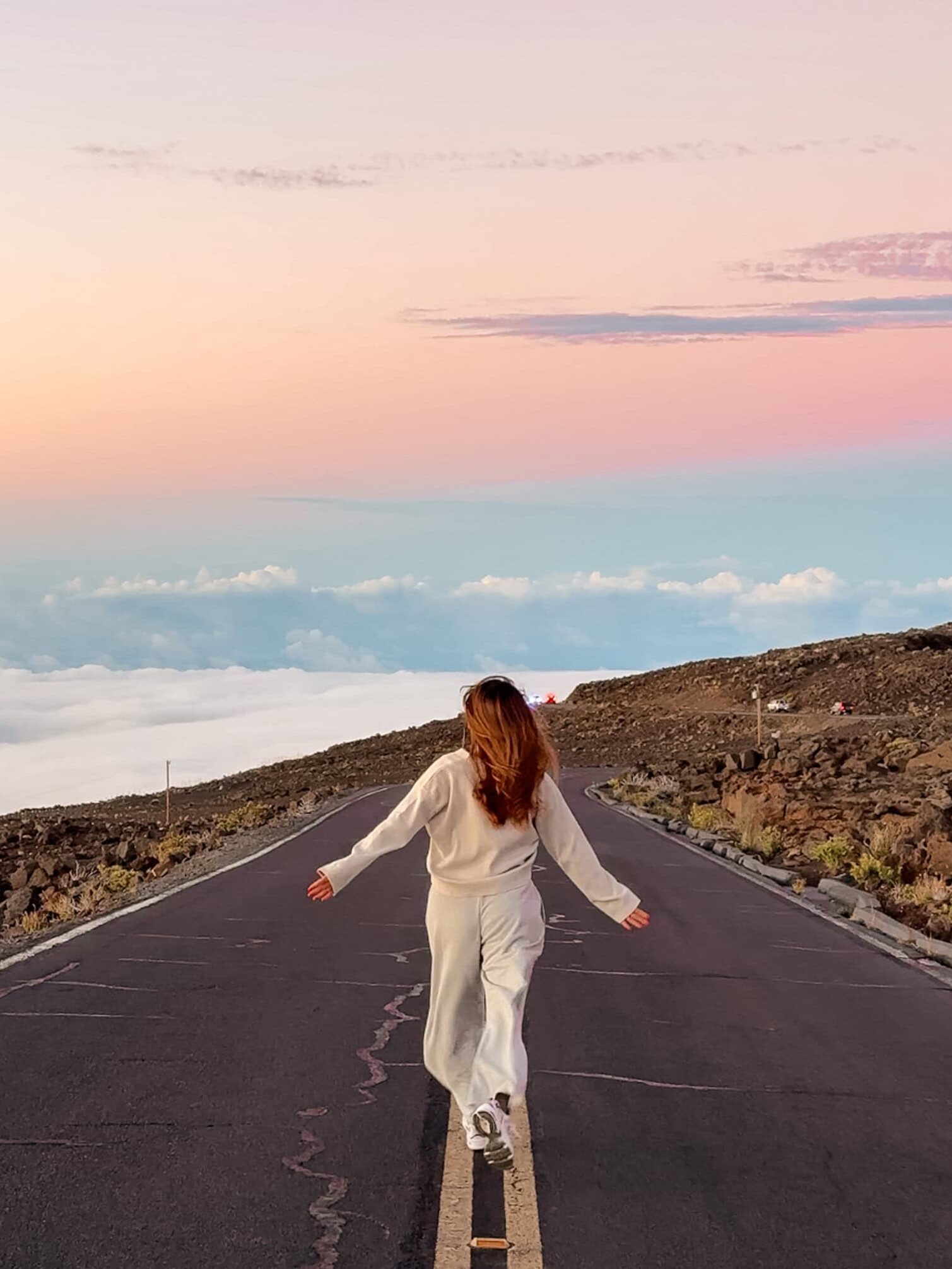 Woman walking along the road above the clouds at Haleakalā National Park during sunset, surrounded by pastel skies and volcanic landscape