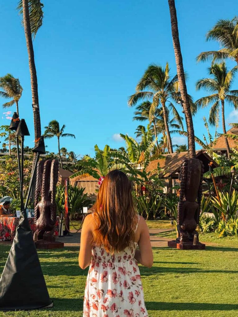Woman in a floral dress standing on the grass surrounded by palm trees and tiki statues during golden hour at Maui Nui Luau in Lahaina