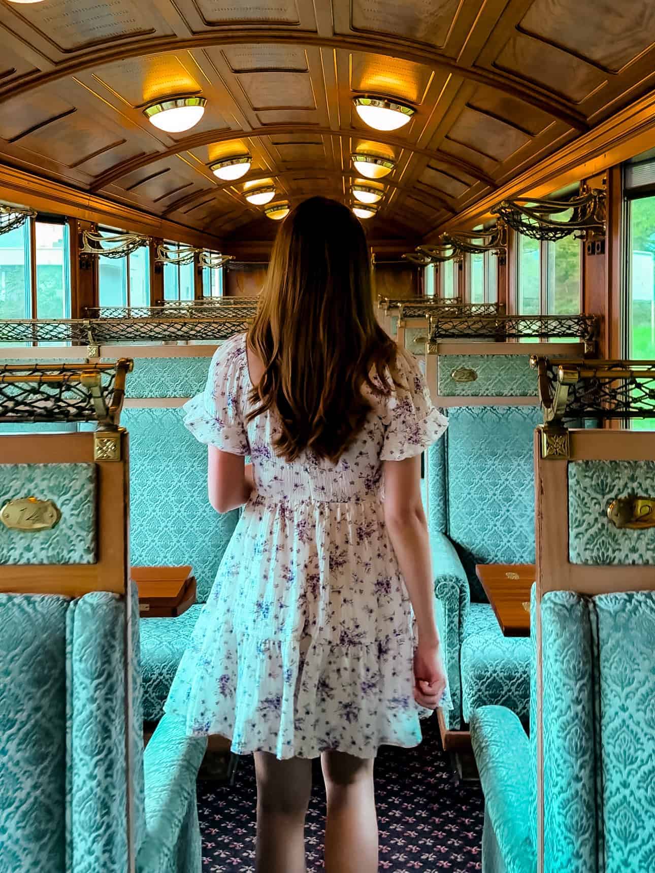 A woman in a light floral dress walks down the aisle of the GoldenPass Belle Epoque train, surrounded by luxurious green velvet seats, ornate brass luggage racks, and warm wooden interiors that exude vintage charm.