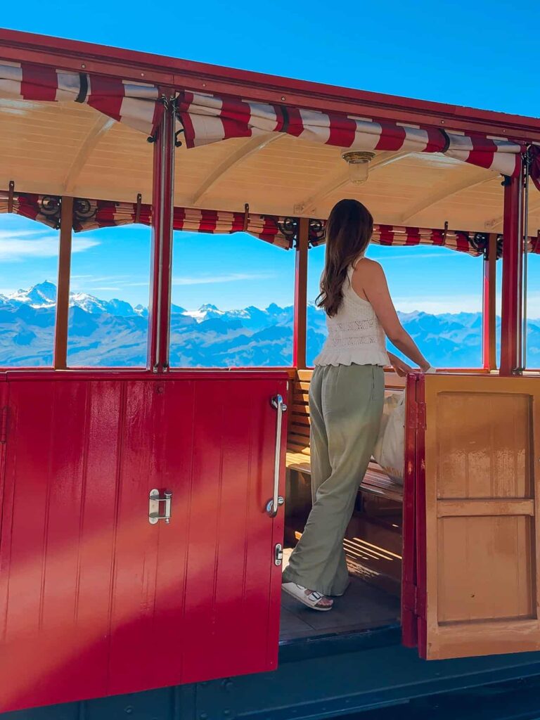 Woman standing on the open carriage of the vintage Brienz Rothorn Bahn train, admiring panoramic views of the Swiss Alps under a clear blue sky.