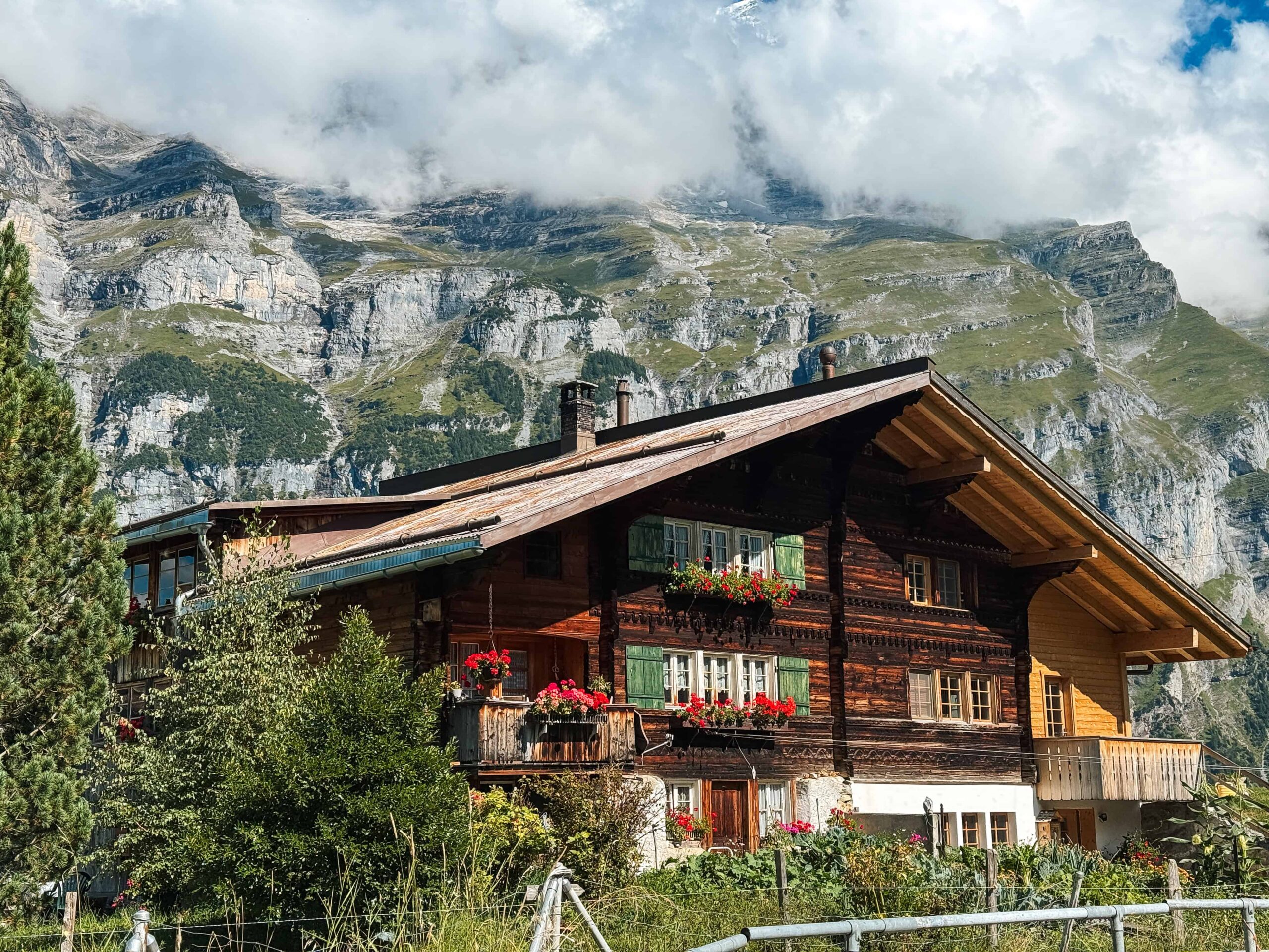 Traditional wooden Swiss chalet adorned with red flower boxes sits in front of dramatic rocky mountain cliffs partially covered in clouds in Gimmelwald, Switzerland which is a must visit in a Switzerland Itinerary