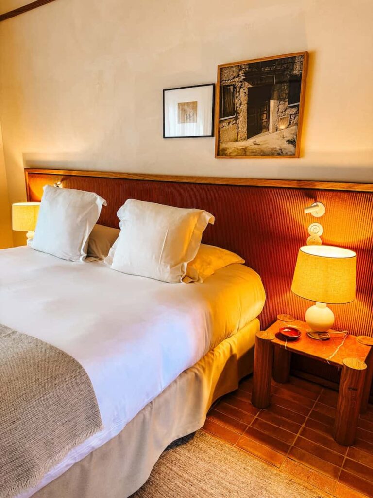 A room at Capelongue hotel with a neatly made bed, warm bedside lamps, and framed artwork above a wooden headboard.