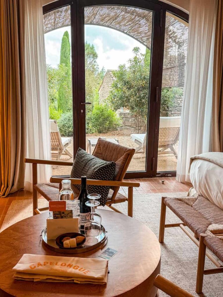 A room at Capelongue hotel with a sitting area with a round wooden table holding a tray with wine, glasses, treats, and a welcome card.