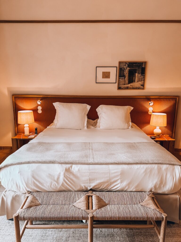 A cozy hotel bed with white linens, soft pillows, warm bedside lamps, and framed artwork above the headboard.