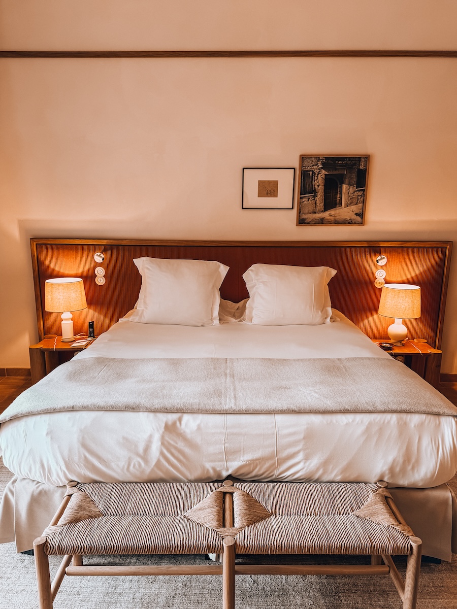 A cozy hotel bed with white linens, soft pillows, warm bedside lamps, and framed artwork above the headboard.