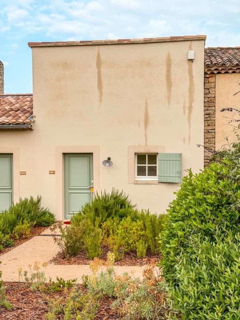 A cream colored cottage façade with a pastel green door and shutter surrounded by rosemary and garden greenery.
