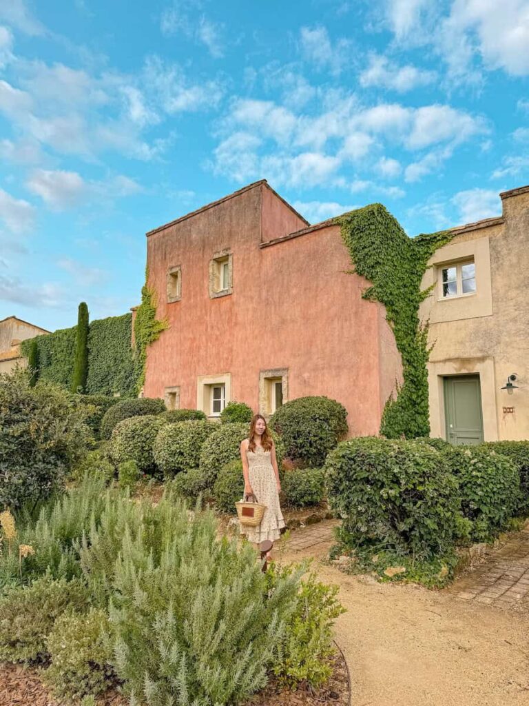 A woman in a floral dress standing in front of an ivy covered terracotta colored villa, surrounded by lush Mediterranean shrubs.