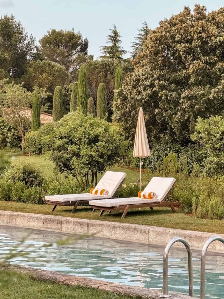 Two white pool loungers with rolled striped towels set beside the garden lined swimming pool.