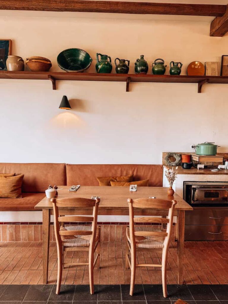 A rustic dining area at Capelongue, with a wooden table, woven chairs, and a bench seat beneath a shelf of ceramic pottery.