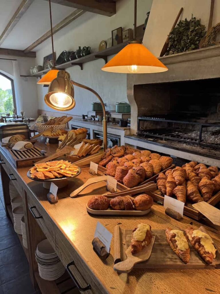 A breakfast table filled with fresh pastries including croissants, breads, and brioche under warm lighting.
