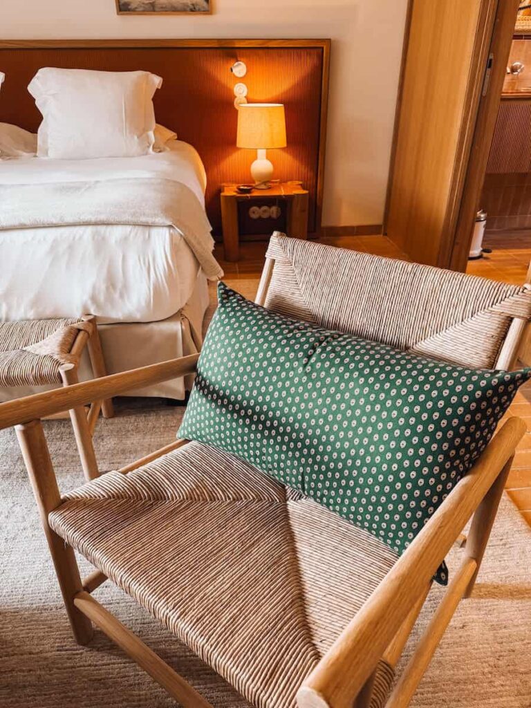A woven lounge chair with a green patterned cushion in a softly lit room beside the bed.