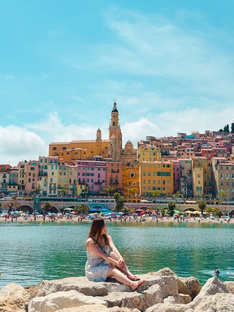 A woman sitting on large rocks by the sea looking across the turquoise water toward Menton’s pastel old town and basilica under a bright blue sky.