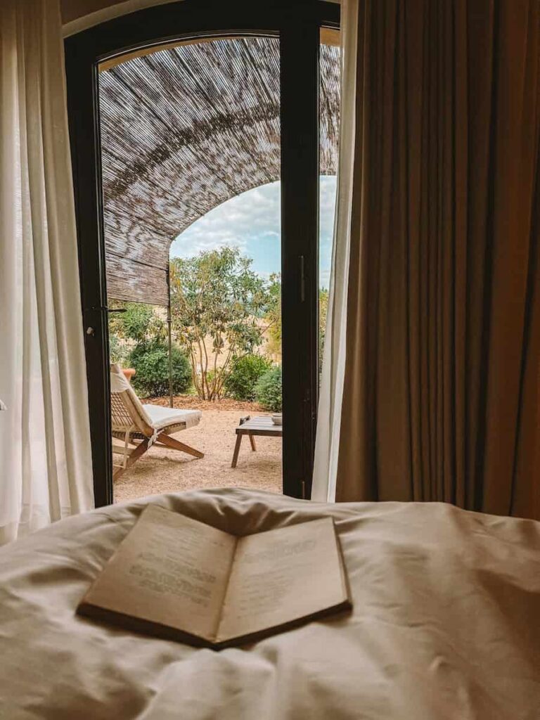 A serene view from the bed with an open book in the foreground, looking out to a terrace and garden through arched glass doors at capelongue hotel. 